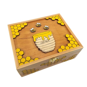 Puede incluir: Una caja de madera con un diseño de tarro de miel y abejas. La caja está decorada con patrones de panal de abeja amarillos y tiene un cierre dorado.