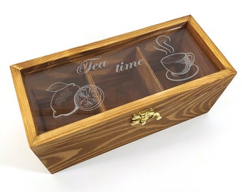 Könnte beinhalten: Eine Holzteebox mit einem durchsichtigen Deckel, der drei Fächer aufweist. Der Deckel ist mit den Worten "Tea time", einer Zitrone und einer Teetasse graviert. Die Box hat einen goldenen Verschluss und eine natürliche Holzoberfläche.