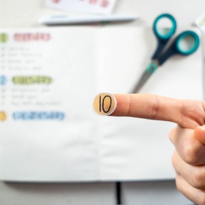 May include: A close-up of a finger holding a round, beige sticker with the number "10" written in black.