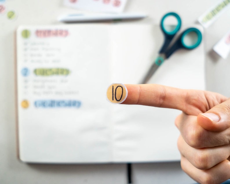 May include: A close-up of a finger holding a round, beige sticker with the number "10" written in black.