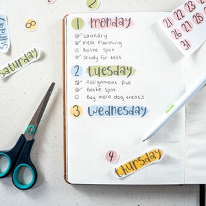 May include: A weekly planner with colorful stickers for each day of the week. The stickers have the days of the week written on them in a handwritten style. The planner is open to the Monday and Tuesday pages, with handwritten to-do lists for each day.