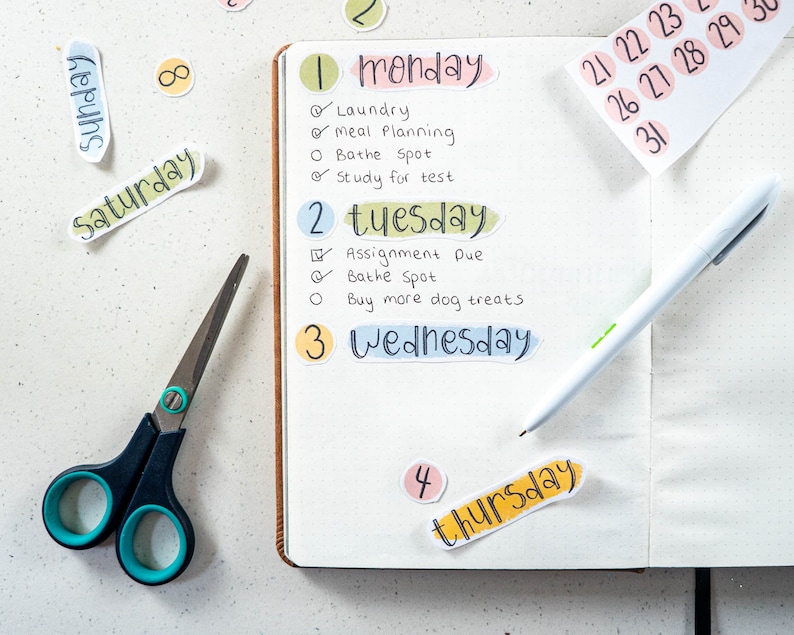 May include: A weekly planner with colorful stickers for each day of the week. The stickers have the days of the week written on them in a handwritten style. The planner is open to the Monday and Tuesday pages, with handwritten to-do lists for each day.