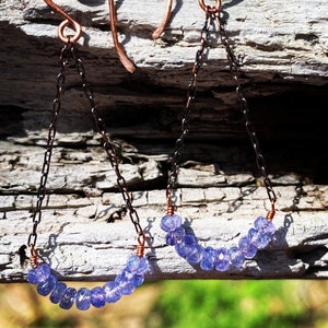May include: A pair of dangle earrings with a copper chain and a row of small, light purple gemstones. The earrings are hanging from a wooden surface.
