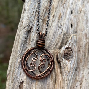 May include: A copper wire necklace with a circular pendant featuring a swirling design. The necklace is hanging on a piece of driftwood.
