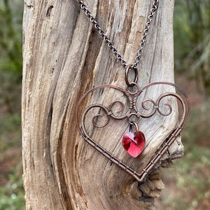 May include: A copper wire heart necklace with a red crystal pendant. The necklace is hanging on a piece of driftwood.