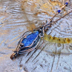 May include: A copper wire wrapped pendant featuring a blue lapis lazuli stone. The pendant is attached to a copper chain with blue beads.