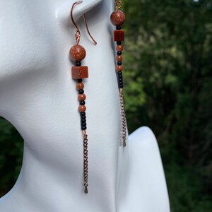 May include: Long dangle earrings with brown and black beads and a gold chain. The beads are round and square.