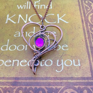 May include: A copper wire heart necklace with a pink gem in the center. The necklace is on a book with the text "Seek and you will find, Knock and the door will be opened to you".