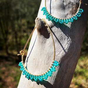 May include: A pair of gold hoop earrings with turquoise blue beads. The earrings are hanging from a wooden surface.