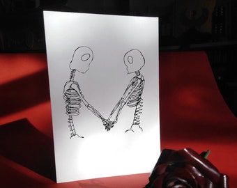 May include: A black and white line drawing of two skeletons holding hands, printed on a white card. The card is sitting on a red surface with a dark red rose.