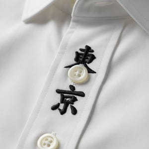 Puede incluir: Primer plano de una camisa blanca con caracteres japoneses bordados en negro. La camisa tiene cuello abotonado y botones blancos. Los caracteres japoneses están encima de los botones.