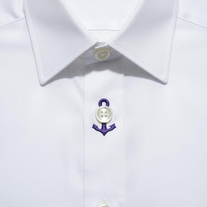 May include: Close-up of a white dress shirt with a purple anchor design embroidered below the collar button. The shirt has a classic collar and a buttoned placket. The anchor is a nautical design.