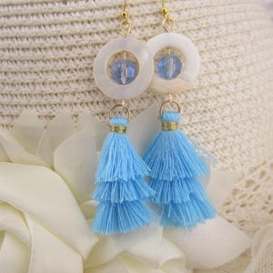 May include: A pair of earrings featuring white circular pendants with blue crystal beads and gold-colored hooks. Below, light blue tiered tassels hang, creating a layered effect. The earrings are set against a soft, textured background.