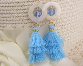 May include: A pair of earrings featuring white circular pendants with blue crystal beads and gold-colored hooks. Below, light blue tiered tassels hang, creating a layered effect. The earrings are set against a soft, textured background.