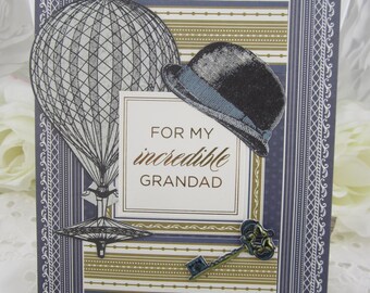 May include: A rectangular greeting card featuring a dark blue and gold striped background with a white central panel reading FOR MY incredible GRANDAD. The card displays a black hot air balloon, a black bowler hat, and a metal key.
