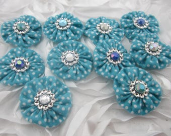 May include: A collection of turquoise fabric flower embellishments with white polka dots. Each flower has a sparkling rhinestone center. The flowers are arranged on a white, textured surface, creating a decorative and eye-catching display.