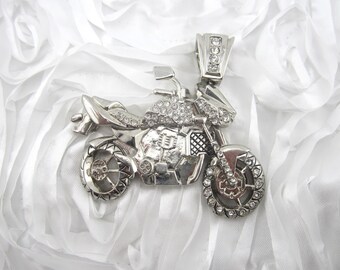 May include: Silver-toned motorcycle pendant with clear crystal accents. The pendant features detailed design elements, including wheels, a seat, and handlebars. The motorcycle is set against a white, textured background.