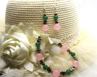 May include: A jewelry set featuring a bracelet and matching earrings. The bracelet and earrings have pink and green beads with silver-colored accents. The jewelry is displayed on a white hat and a white floral arrangement.
