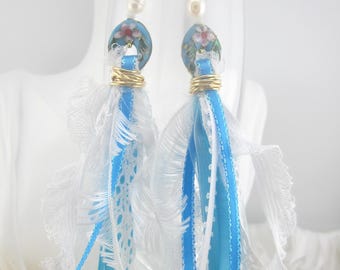 May include: Elegant dangle earrings featuring blue and white ribbons, lace, and floral accents. The earrings have gold-colored hooks and wire, with a pearl bead and a floral design on the top. The earrings are a unique accessory.