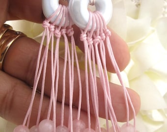 May include: Dangle earrings featuring white ceramic donut-shaped beads, connected to pink cord tassels. Each tassel ends with a pink gemstone bead. The earrings have silver-colored ear wires.