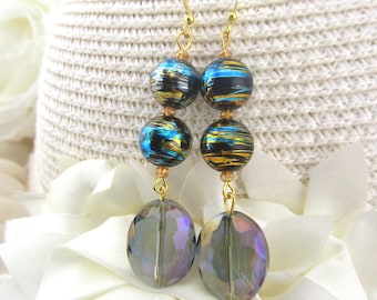 May include: Dangle earrings with gold-colored hooks. Each earring features two striped blue and gold beads, and a large, faceted, oval-shaped, iridescent bead. The earrings are displayed on a white background.