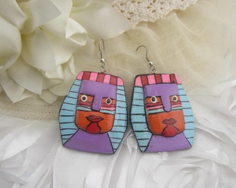 May include: A pair of handmade, colorful earrings. Each earring features a stylized face with a purple head, orange face, and red lips. The design includes blue stripes, pink and red stripes, and silver earring hooks. The earrings are set against a white background.