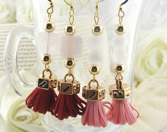 May include: Four pairs of dangle earrings with gold-colored hooks and accents. Each earring features a combination of beads and a small tassel. The tassels are in shades of red and pink, with a clear glass cup in the background.