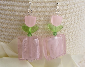 May include: A pair of dangle earrings featuring pink tulip-shaped beads with green leaf accents and square pink glass blocks. The earrings have silver-colored hooks and are set against a cream-colored background.
