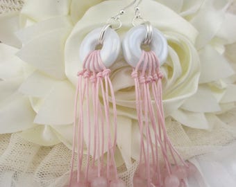 Puede incluir: Pendientes hechos a mano con cuentas blancas en forma de rosquilla, macramé rosa y cuentas de piedras preciosas rosas. Los pendientes cuelgan de ganchos plateados. Se presentan sobre un fondo floral blanco.