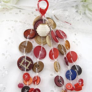 May include: A decorative hanging ornament featuring a red plastic lobster clasp attached to multiple white strings. Strung along these threads are various round buttons in shades of red, brown, tan, blue, and burgundy, along with a central white bead.