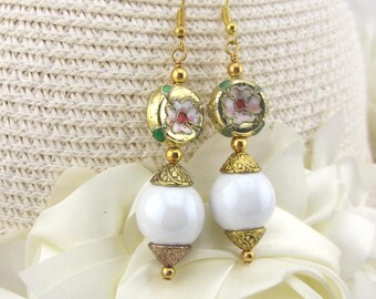 May include: A pair of dangle earrings with gold-colored hooks. Each earring features a white, round bead, topped with a gold-colored filigree and a floral design in pink, red, and green. The earrings are set against a white background.