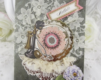 May include: A rectangular greeting card featuring a dark gray background with a white lace overlay. The card displays a vintage black telephone, a pink and brown floral paper cutout, a purple flower, and a pink banner with the text Hello.