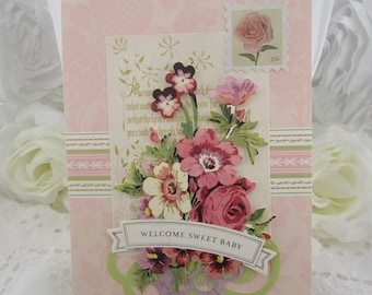 May include: A greeting card with a light pink background featuring a floral bouquet, a postage stamp with a rose, and a banner with the text WELCOME SWEET BABY. The card includes decorative green accents and script text on a cream panel.