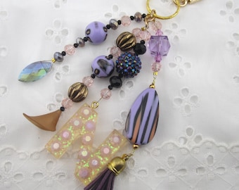 May include: A keychain featuring a gold-tone lobster clasp and ring, adorned with a variety of beads including purple, black, and metallic tones. A letter M charm with pink glitter accents and a small purple tassel are attached to the bead strands.
