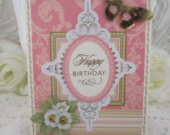 May include: A rectangular greeting card with a pink damask background, featuring a central oval plaque with the text Happy BIRTHDAY. The card is decorated with a brown butterfly cutout at the top right and three white flowers with yellow centers at the bottom left.