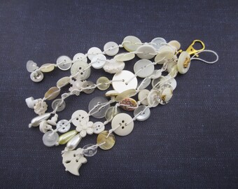 May include: A decorative tassel charm composed of numerous white and cream-colored buttons of varying sizes and shapes, strung together on white cord. The cluster features a gold-toned metal lobster clasp at the top for attachment.