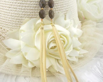 May include: Gold-tone earrings with brown stone beads and long, beige suede fringe. The earrings are displayed on a white hat with a floral design, creating a bohemian aesthetic. The earrings are a stylish accessory.