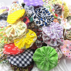 May include: A collection of colorful fabric yo-yos in various patterns and colors, including floral, polka dots, and geometric designs. The yo-yos are in shades of yellow, green, red, purple, and blue. These fabric circles are a craft supply.