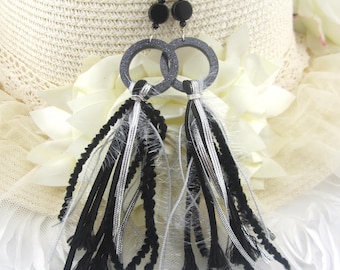 May include: A pair of dangle earrings featuring black and silver tassels. The earrings have gray circular hoops and black bead accents. The tassels are made of black and silver strands. The earrings are displayed on a white background.