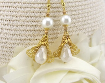May include: A pair of elegant gold-toned earrings with a pearl and teardrop design. Each earring features a large white pearl, smaller gold beads, and a delicate filigree bell shape holding a pear-shaped white gem. The earrings hang from gold-colored hooks.