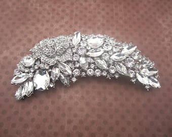 May include: A silver-toned, curved hair accessory adorned with numerous clear, sparkling rhinestones. The design features a central floral motif and a variety of oval and teardrop-shaped stones, creating a glamorous, eye-catching piece.
