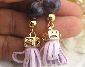 May include: A pair of dangle earrings with gold-colored hooks, beads, and square accents. Each earring features a dark purple bead, a gold bead, and a light purple tassel. The earrings are displayed against a white background.
