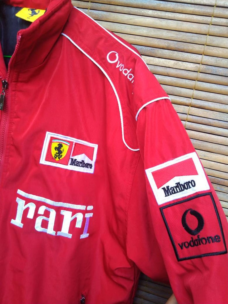 Puede incluir: Chaqueta roja Ferrari con ribetes blancos y logotipos bordados para Marlboro, Vodafone y Ovodafone. La chaqueta tambi&eacute;n tiene la palabra "Ferrari" bordada en la parte delantera.