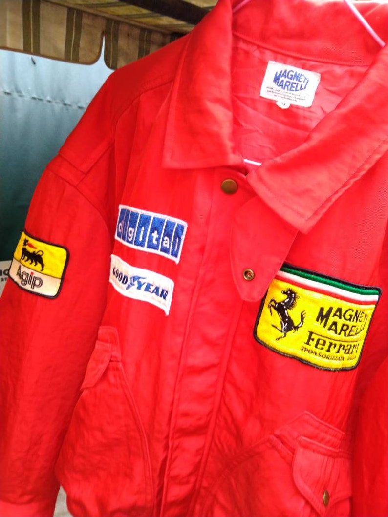 Op de afbeelding: Felrood jack met meerdere patches, waaronder "Agip", "digital" en "Goodyear". De jas heeft ook een patch met een zwart paardenlogo en de woorden "MAGNETI MARELLI Ferrari". De jas heeft een kraag en twee voorzakken.
