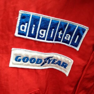 Op de afbeelding: Close-up van een rode overall met twee geborduurde patches. De bovenste patch toont het woord "DIGITAL" in witte letters op een blauwe achtergrond. De onderste patch zegt "GOODYEAR" in blauwe letters op een witte achtergrond.