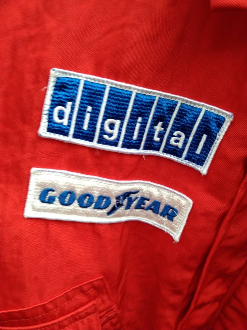 Op de afbeelding: Close-up van een rode overall met twee geborduurde patches. De bovenste patch toont het woord "DIGITAL" in witte letters op een blauwe achtergrond. De onderste patch zegt "GOODYEAR" in blauwe letters op een witte achtergrond.