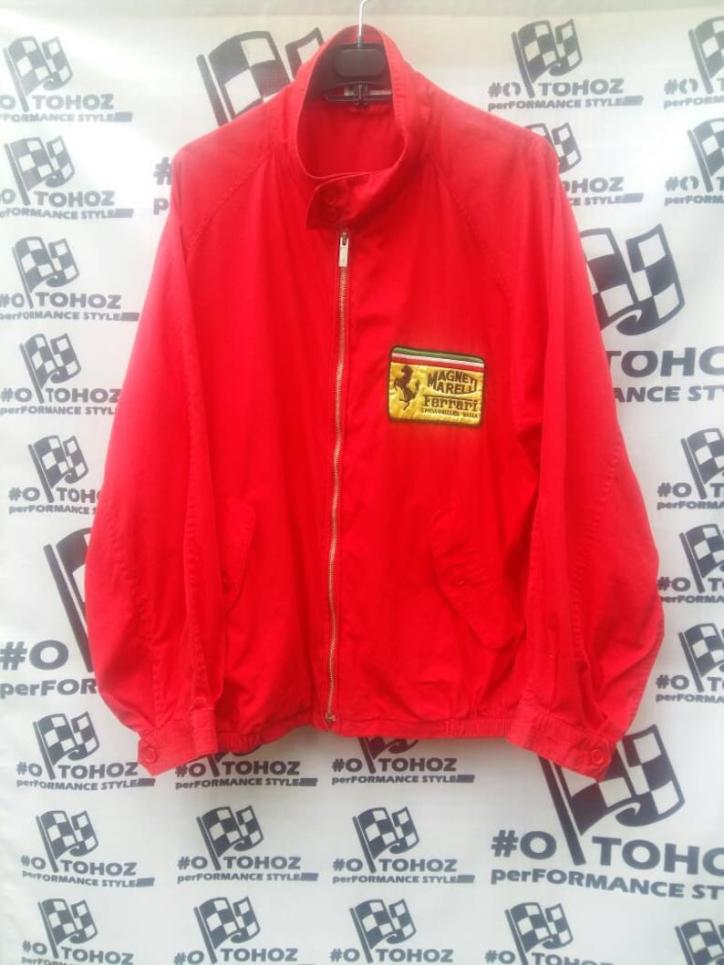K&ouml;nnte beinhalten: Eine rote Rei&szlig;verschlussjacke mit einem Ferrari-Logo-Patch. Der Patch ist mit den Worten "Magnet Marelli Ferrari" und "Scuderia Ferrari" bestickt.