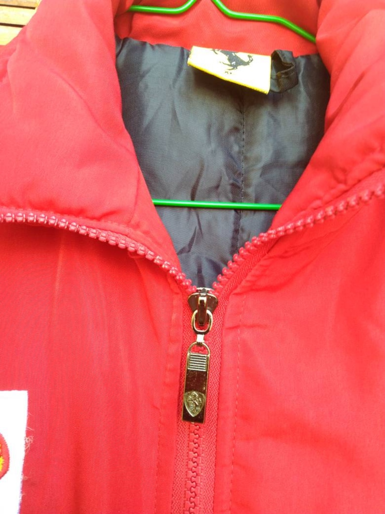 Puede incluir: Chaqueta roja con cremallera, con forro negro y un tirador de cremallera dorado con el logotipo de Ferrari. La chaqueta tiene una etiqueta amarilla con el logotipo de Ferrari en el interior del cuello.