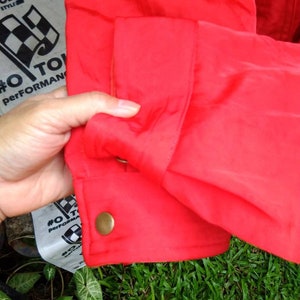 Op de afbeelding: Close-up van een felrode jas mouw. De mouw heeft een manchet met knoop en een goudkleurige knoop. De stof heeft een lichte glans. De achtergrond omvat groen gras en een gedeeltelijk zichtbaar label met tekst en een geblokte vlag.