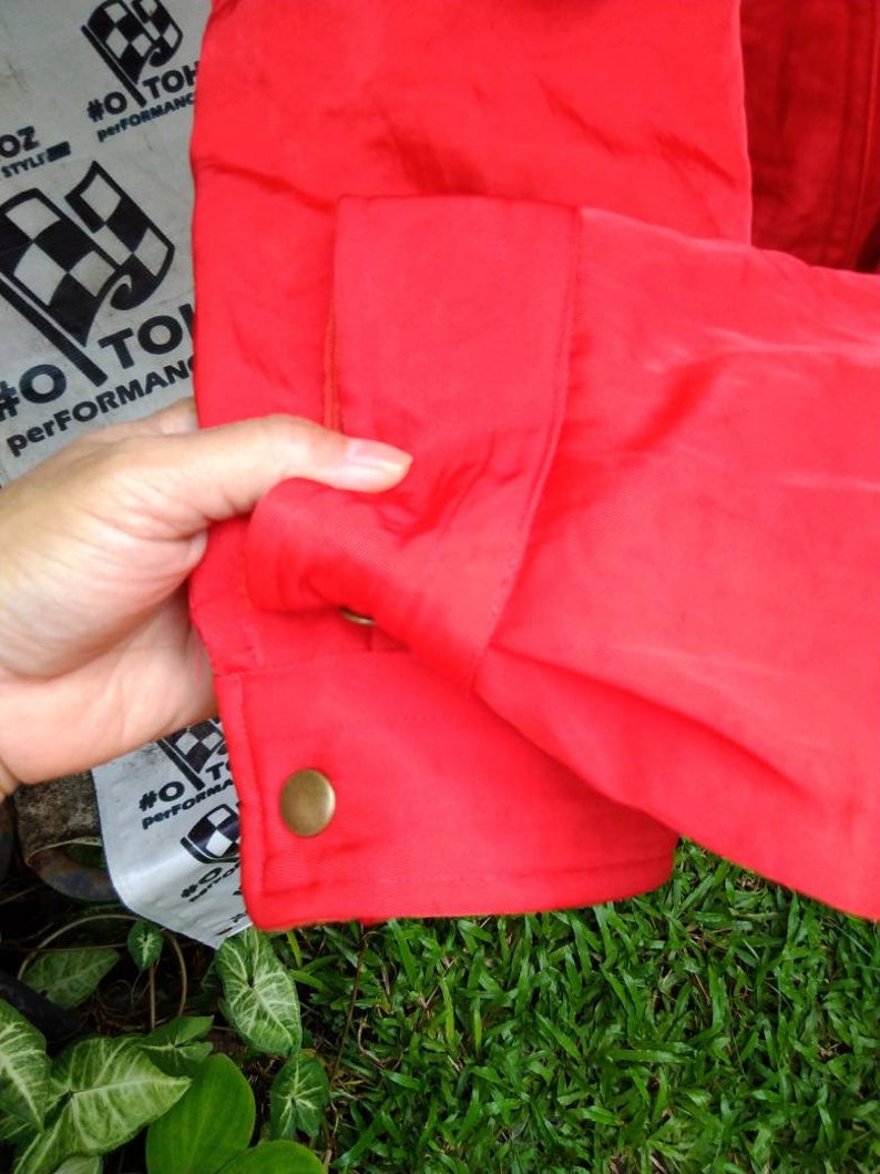 Op de afbeelding: Close-up van een felrode jas mouw. De mouw heeft een manchet met knoop en een goudkleurige knoop. De stof heeft een lichte glans. De achtergrond omvat groen gras en een gedeeltelijk zichtbaar label met tekst en een geblokte vlag.